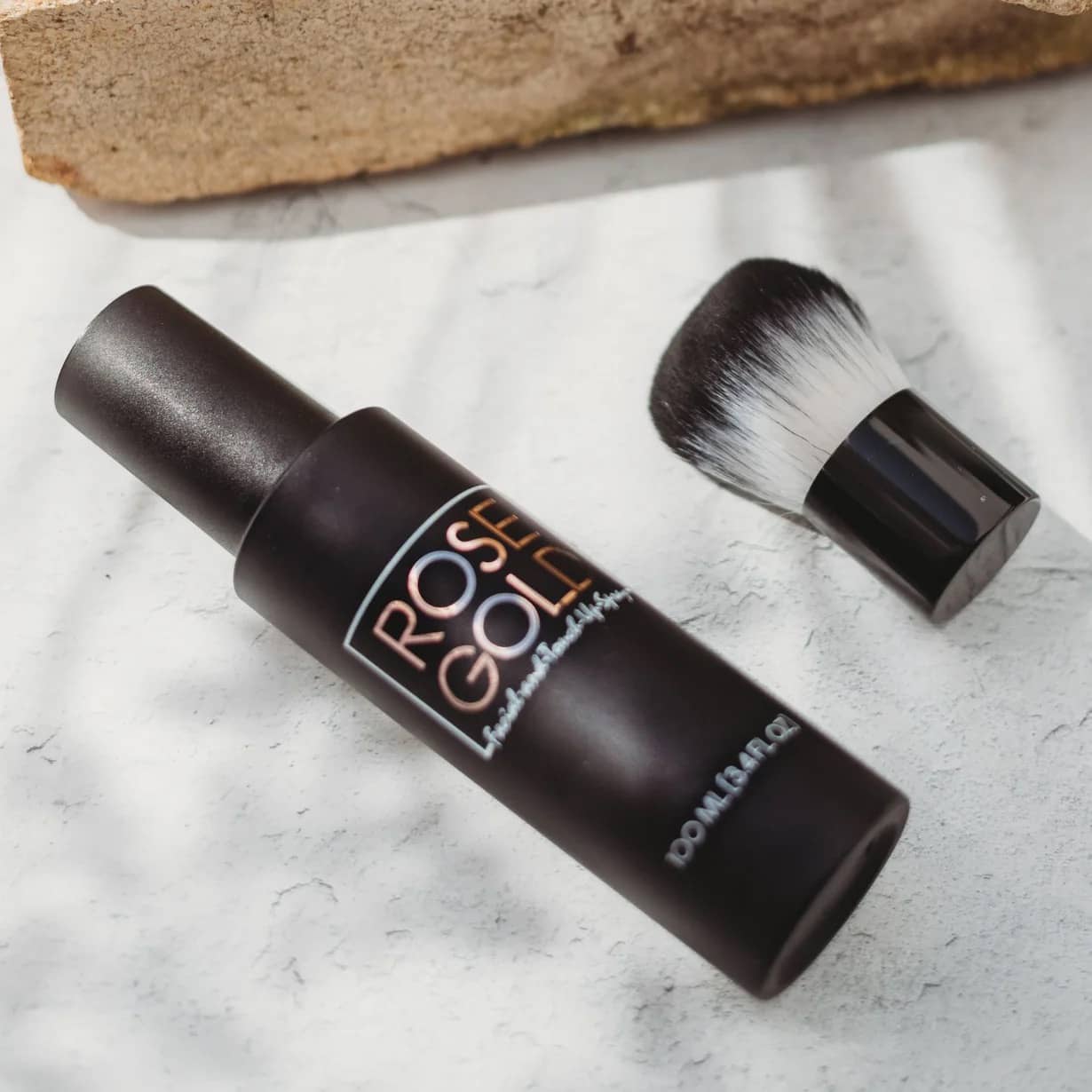 Rose Gold Sunless facial tanning mist bottle with tanning brush on light stone background flat lay